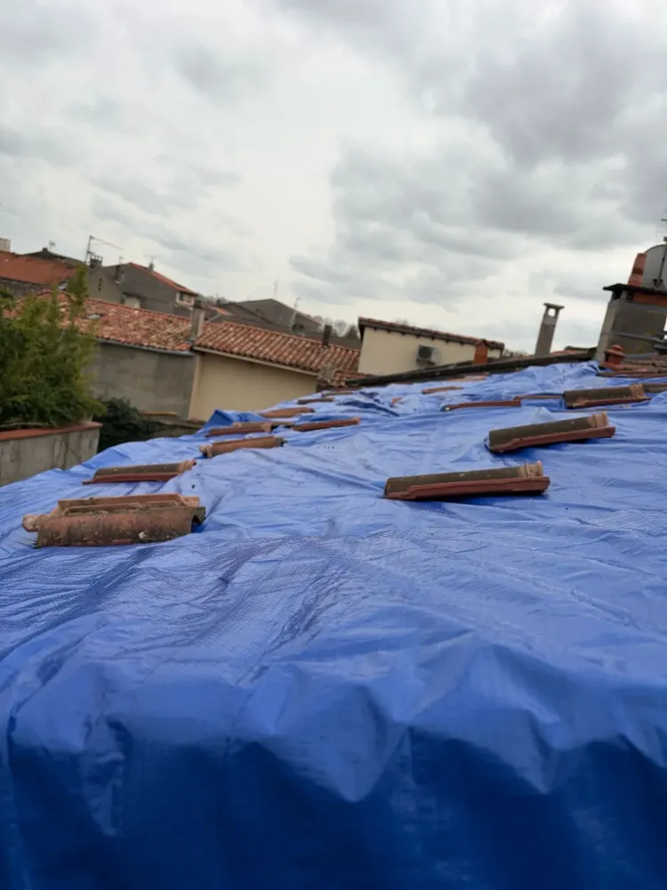 Détail d'une section de toiture couverte par une bâche de protection bleue temporaire, maintenue par des tuiles individuelles éparpillées