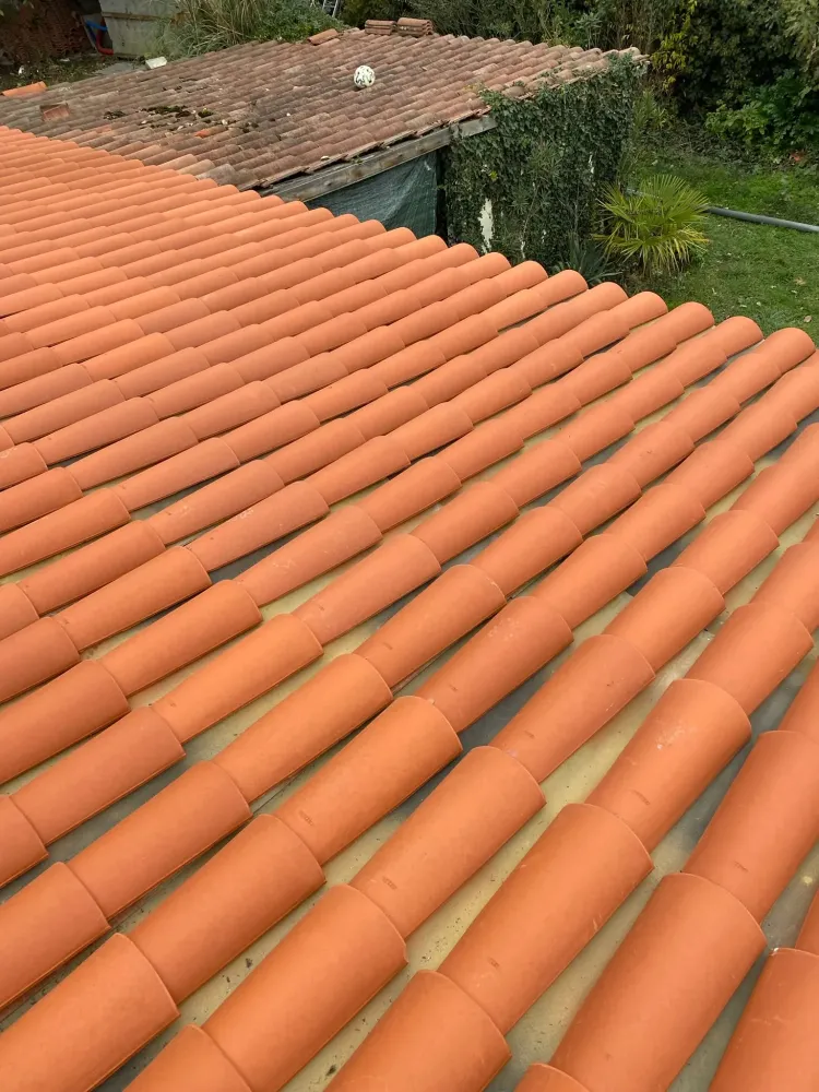 Alignement régulier de tuiles neuves posées sur des plaques ondulées, avec l'ancienne toiture d'une annexe visible en arrière-plan